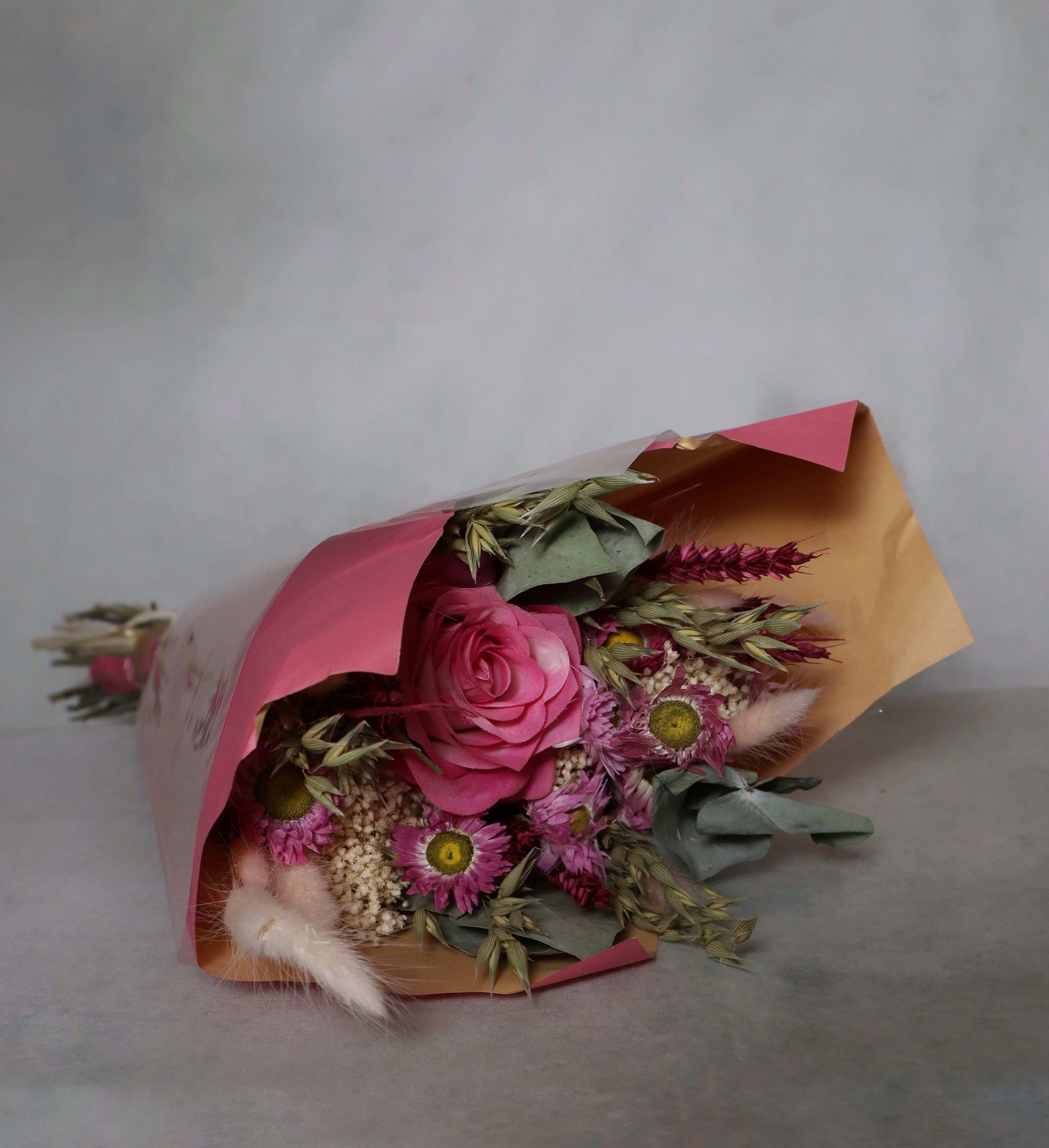 Dried pink flower bouquet wrapped in brown and pink paper on a grey background.