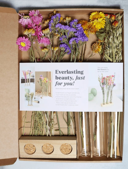 Open box containing dried flowers in pink, purple, yellow, and orange with glass tubes and a wooden stand on a white background.