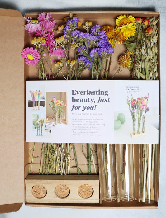Open box containing dried flowers in pink, purple, yellow, and orange with glass tubes and a wooden stand on a white background.