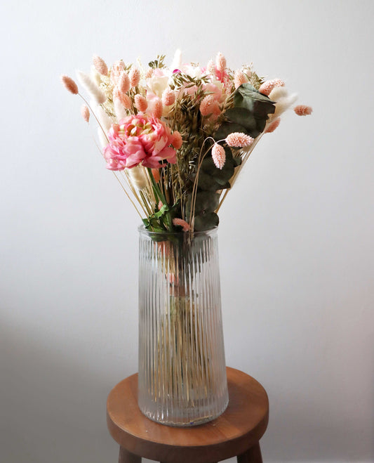 A tall glass vase on a wooden stool, with a bouquet of dried flowers. The arrangement includes pink and white blooms, peach and beige dried grasses, and green foliage, against a light grey background.