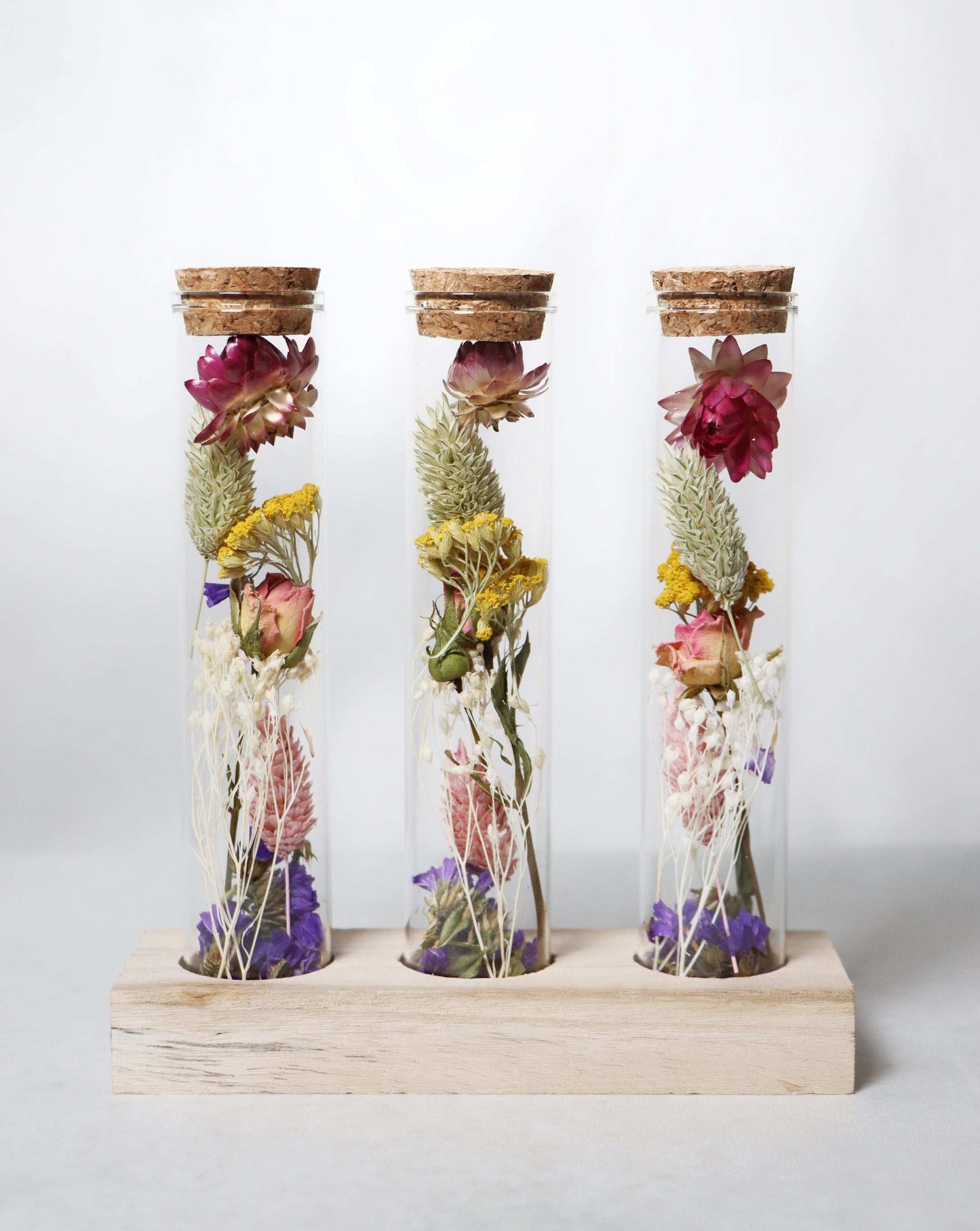 Three glass vials with cork tops containing dried flowers in pink, yellow, and purple on a wooden stand against a white background.