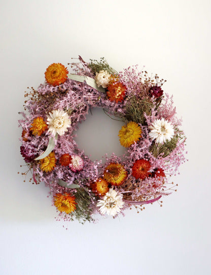 A circular wreath made of dried flowers, featuring a mix of pink, orange, red, and white blooms displayed against a plain white background.
