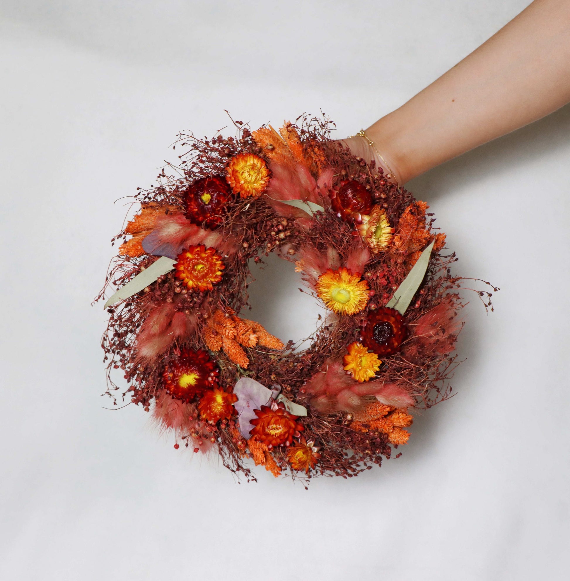 A circular wreath made of dried flowers, featuring a vibrant mix of red, orange, and yellow blooms, along with green foliage accents. The wreath is held in a person's hand against a white background.