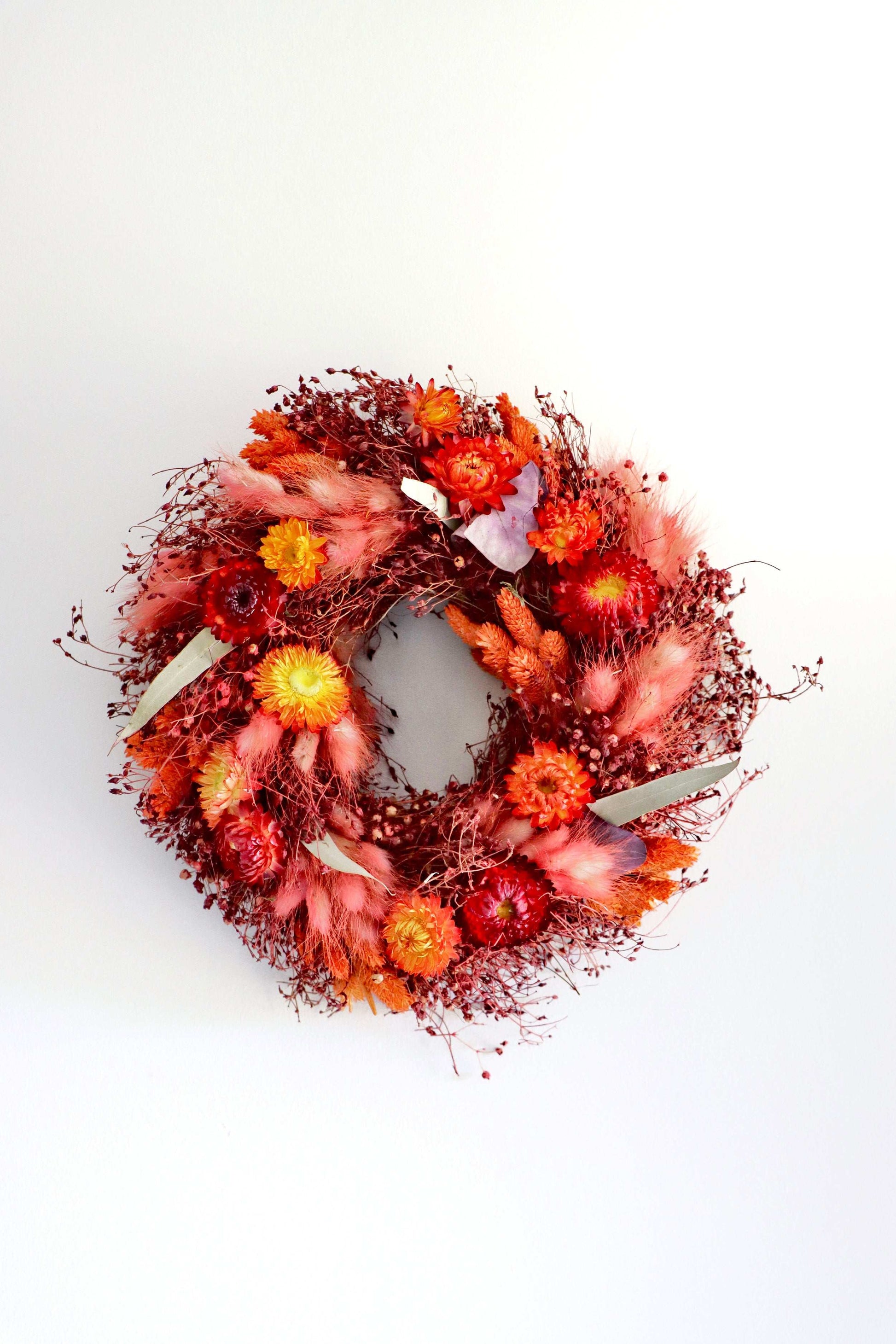 A circular wreath made of dried flowers, featuring a vibrant mix of red, orange, and pink blooms, along with delicate purple foliage accents. The wreath is displayed against a plain white background.