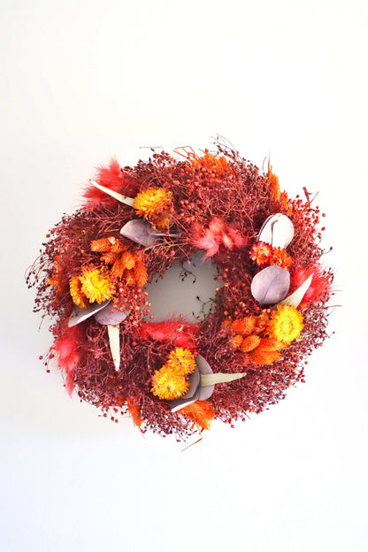 A circular wreath made of dried flowers, featuring a rich mix of red, orange, and yellow blooms, along with purple foliage accents and delicate dried grasses. The wreath is against a plain white background.
