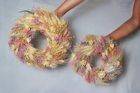 Two circular wreaths made of dried flowers and foliage, featuring pink, yellow, and white blooms, along with tan grasses and green leaves. The wreaths are held in a person's hands against a grey background.