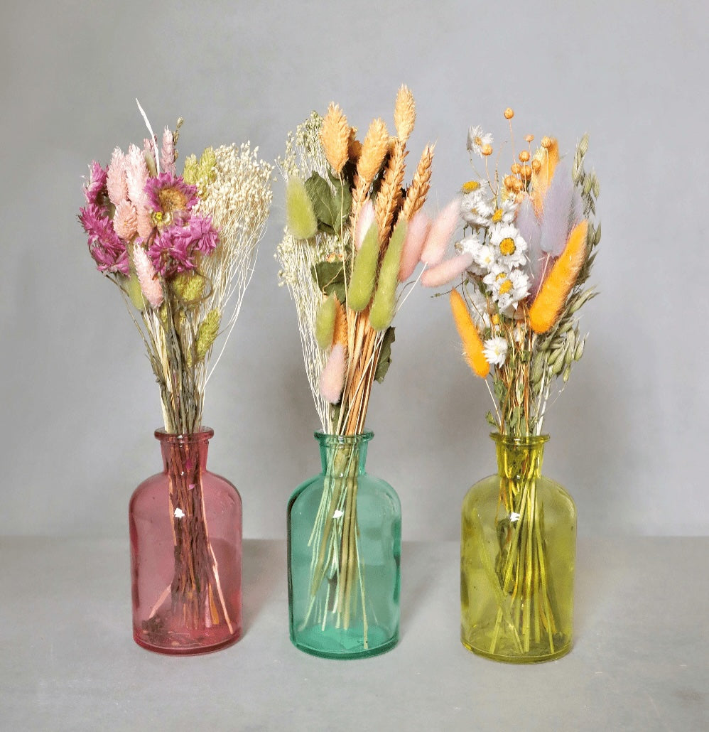 Three dried flower bouquets in pink, blue and yellow glass vases on a grey background.