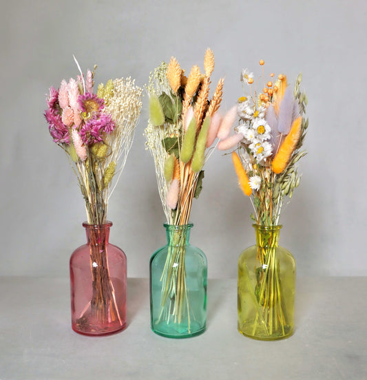 Three dried flower bouquets in pink, blue and yellow glass vases on a grey background. 