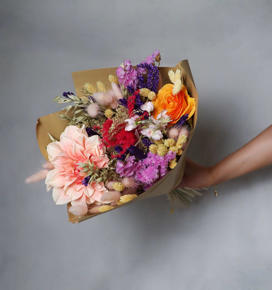 A vibrant bouquet of dried flowers wrapped in brown paper, featuring a pink dahlia, orange rose, purple statice, and dried grasses. Held by a hand against a gray background.