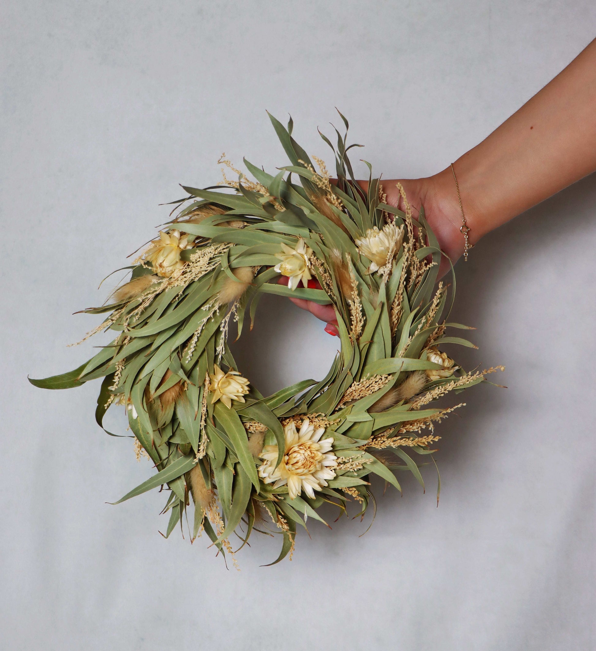 A circular wreath made of dried flowers and foliage, featuring light yellow blooms and green leaves. The wreath is held in a person's hand against a plain grey background.