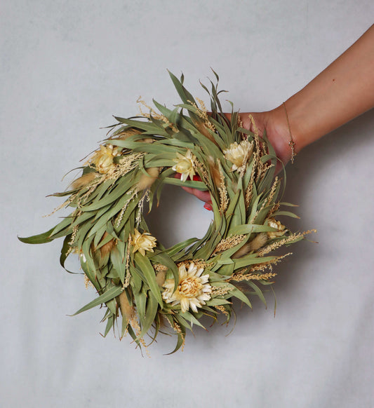 A circular wreath made of dried flowers and foliage, featuring light yellow blooms and green leaves. The wreath is held in a person's hand against a plain grey background.