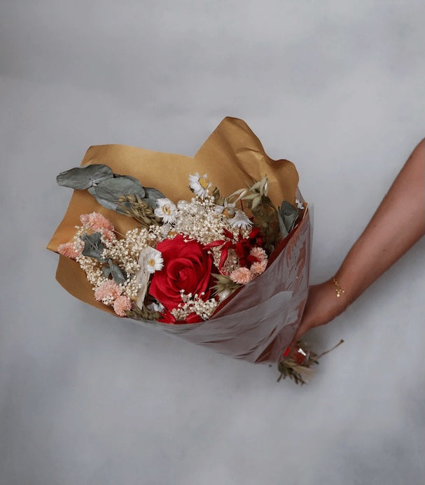Hand holding a bouquet of dried flowers with pink flowers and red roses, eucalyptus, and baby’s breath wrapped in brown paper against a grey background.