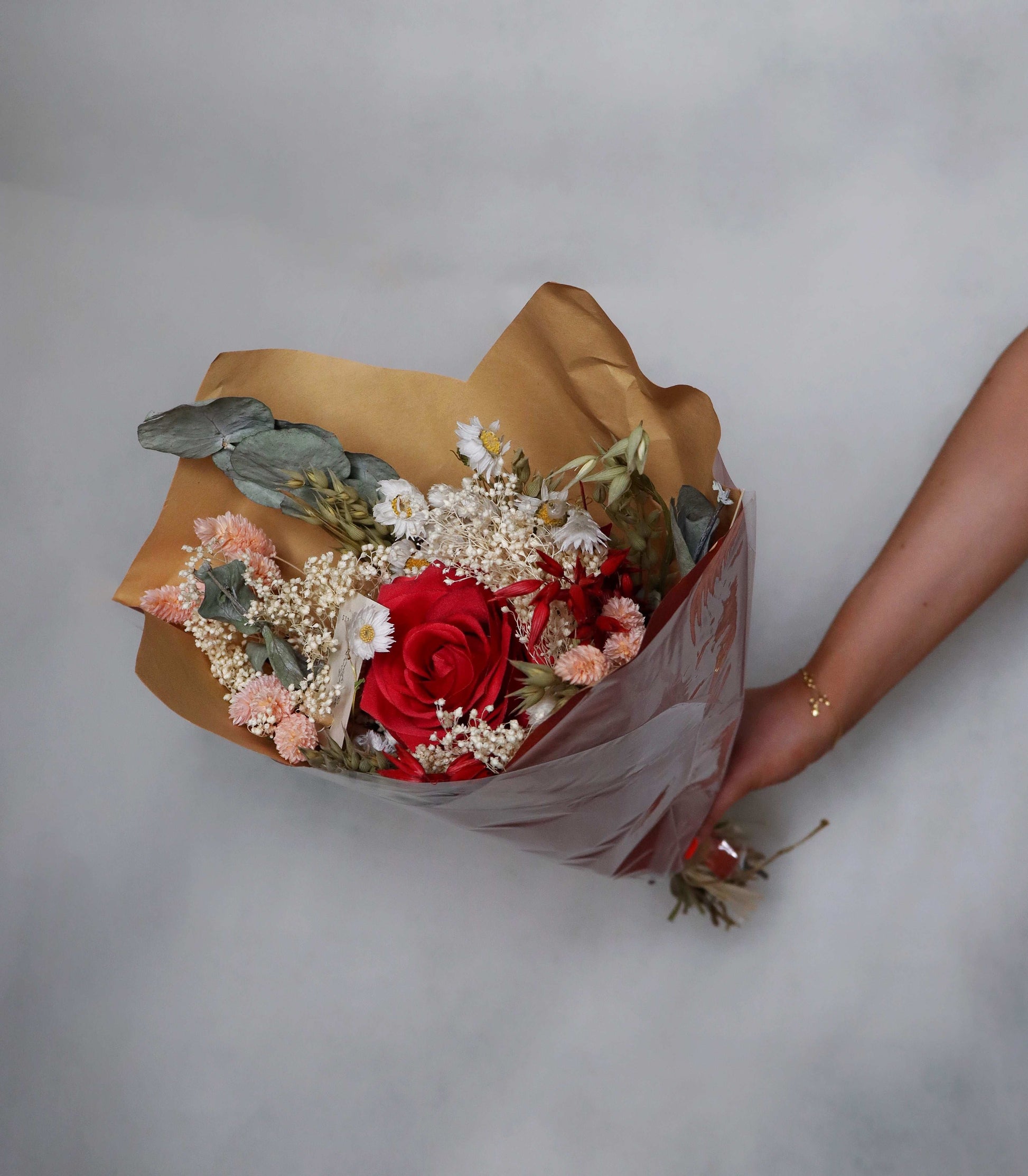 Hand holding a bouquet of dried flowers with pink flowers and red roses, eucalyptus, and baby’s breath wrapped in brown paper against a grey background.