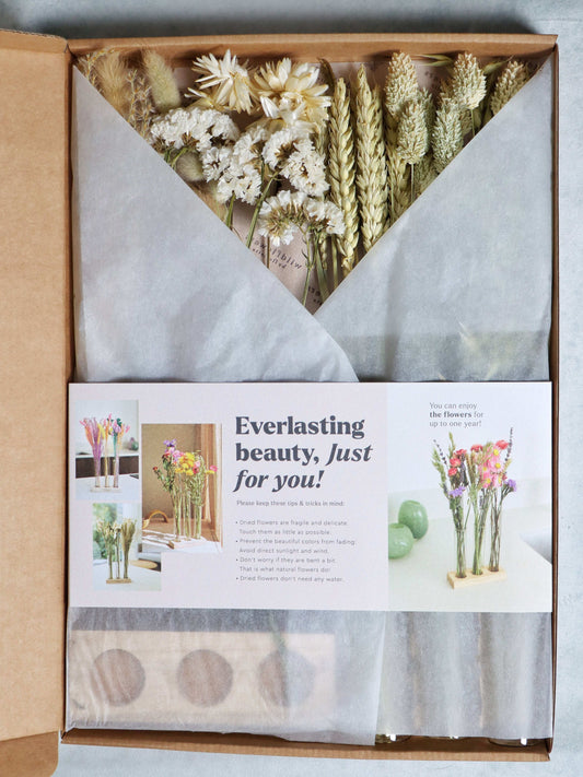 Three glass tubes with dried white and beige flowers, pampas grass, and wheat stalks on a wooden stand against a grey background.