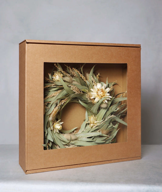 A cardboard box with a windowed front, displaying a circular dried flower wreath inside. The wreath features light yellow blooms and green foliage, set against a grey background.