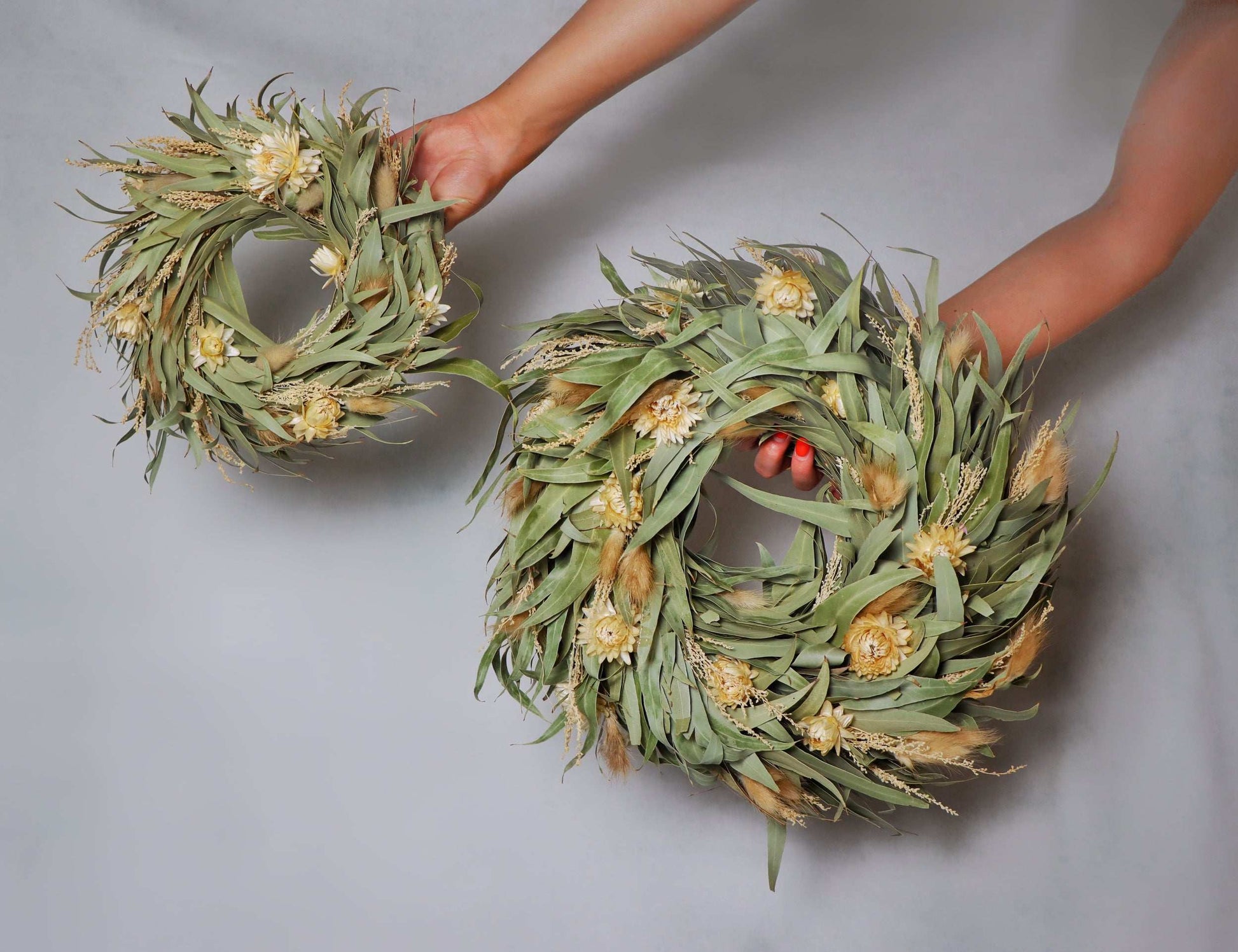 Two circular wreaths made of dried flowers and foliage, featuring light yellow blooms and green leaves. The wreaths are held in a person's hands against a plain grey background.