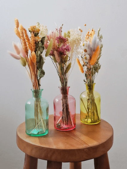 Three dried flower bouquets in blue, pink and yellow glass vases on a wooden stool with a white background.