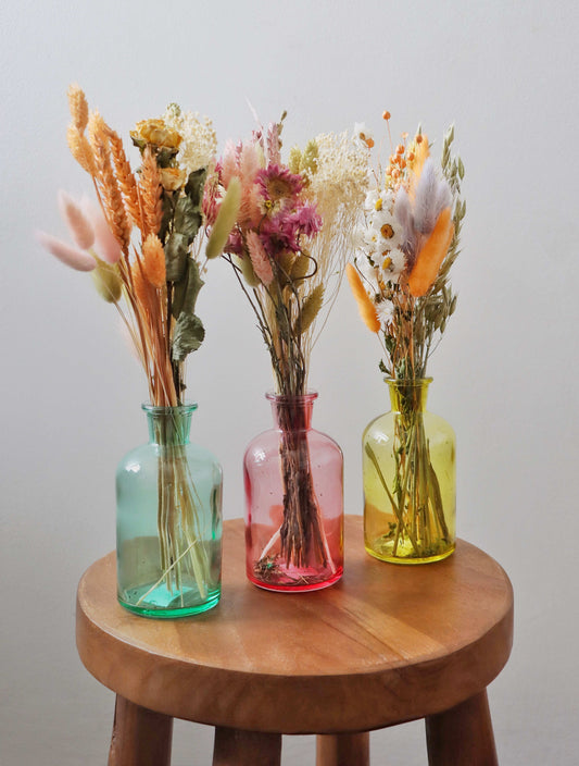 Three dried flower bouquets in blue, pink and yellow glass vases on a wooden stool with a white background. 