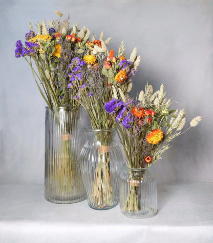 Three glass vases of varying sizes, each filled with dried flowers including purple statice, yellow and orange strawflowers, and dried grasses against a grey background.