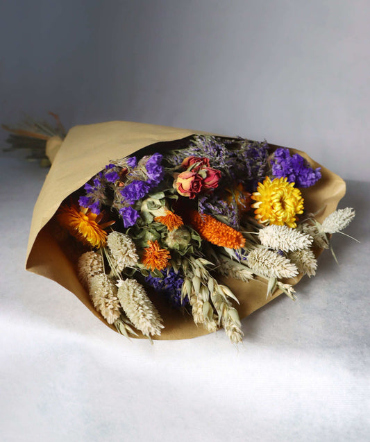A colourful bouquet of dried flowers wrapped in brown paper, featuring purple statice, yellow strawflowers, orange celosia, and dried grasses on a white surface.