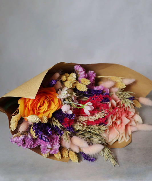 A colourful bouquet of dried flowers wrapped in brown paper, featuring orange, pink, purple, and yellow blooms, along with beige and green stems, set against a grey background.